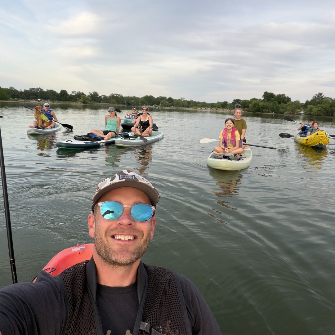 On the water at one of the community paddle gatherings: a selfie with a group of paddleboarders and a yellow kayak spread across the lake behind